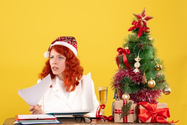 Vooraanzicht vrouwelijke arts zittend achter tafel met documenten op gele achtergrond met kerstboom en geschenkdozen