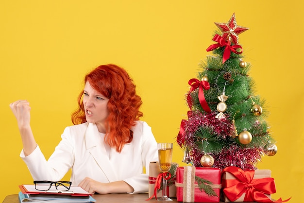 Vooraanzicht vrouwelijke arts zittend achter tafel met kerstcadeautjes bedreigend op gele achtergrond