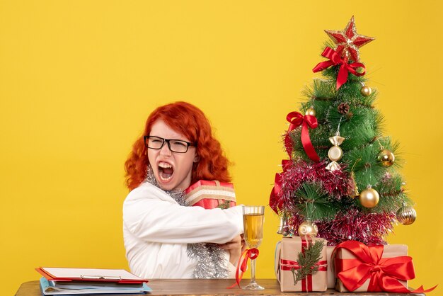 Vooraanzicht vrouwelijke arts zittend achter tafel met kerstcadeautjes op gele achtergrond