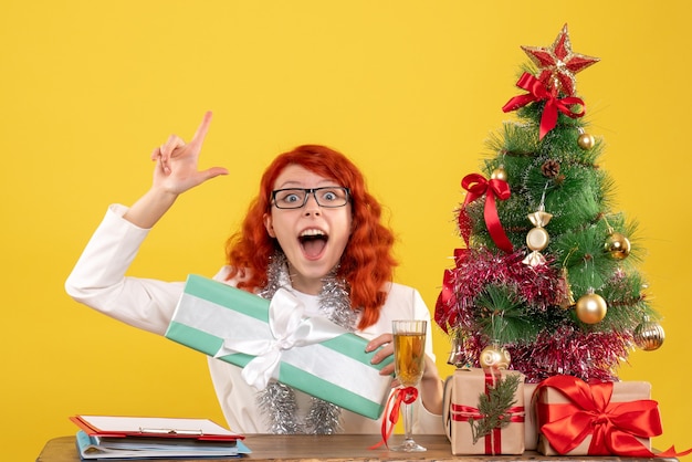 Vooraanzicht vrouwelijke arts zittend achter tafel met kerstcadeautjes op gele achtergrond