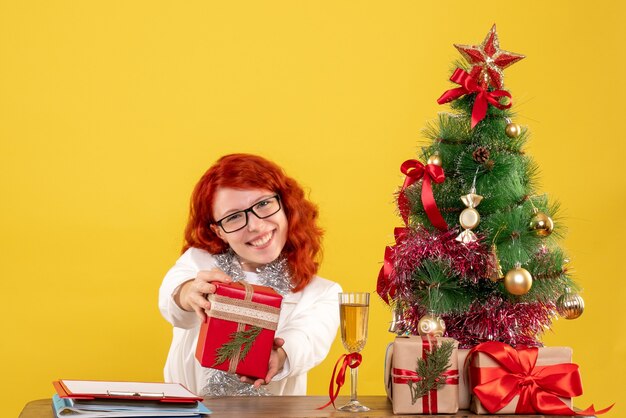 Vooraanzicht vrouwelijke arts zittend achter tafel met kerstcadeautjes op gele achtergrond