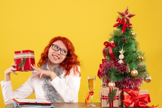 Vooraanzicht vrouwelijke arts zittend achter tafel met kerstcadeautjes op gele achtergrond