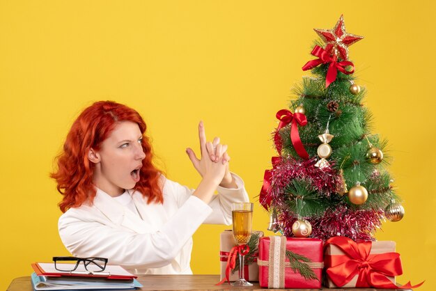 Vooraanzicht vrouwelijke arts zittend achter tafel met kerstcadeautjes op gele achtergrond