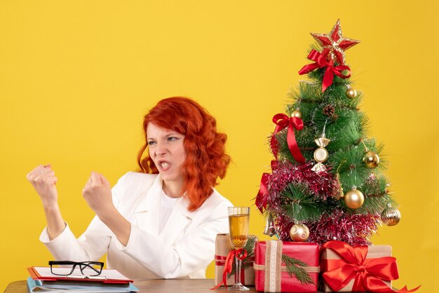 Vooraanzicht vrouwelijke arts zittend achter tafel met kerstcadeautjes op gele achtergrond