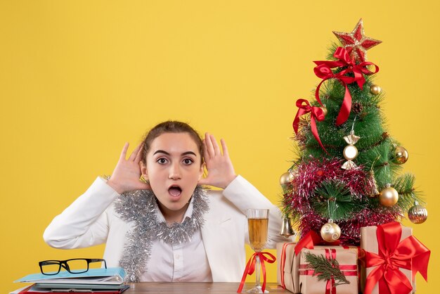 Vooraanzicht vrouwelijke arts zittend achter tafel op gele achtergrond met kerstboom en geschenkdozen