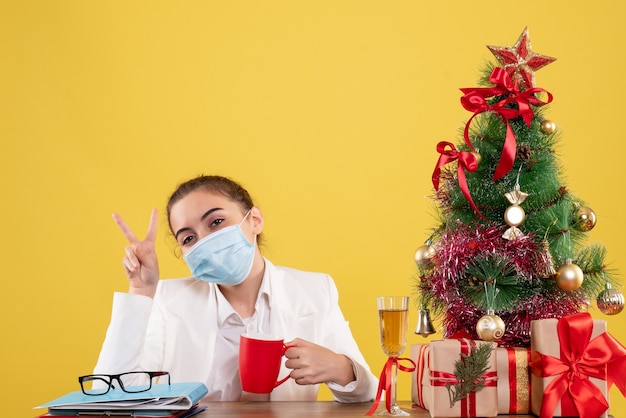 Vooraanzicht vrouwelijke arts zittend in beschermend masker op gele achtergrond met kerstboom en geschenkdozen