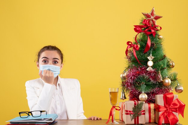 Vooraanzicht vrouwelijke arts zittend in beschermend masker op gele achtergrond met kerstboom en geschenkdozen