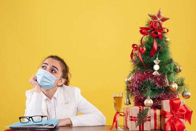 Vooraanzicht vrouwelijke arts zittend in steriel masker beklemtoond op gele achtergrond met kerstboom en geschenkdozen