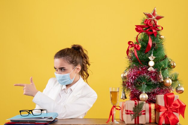 Vooraanzicht vrouwelijke arts zittend in steriel masker op gele achtergrond met kerstboom en geschenkdozen