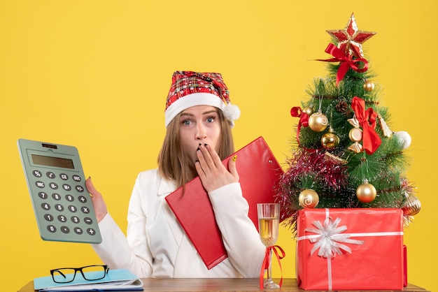 Vooraanzicht vrouwelijke arts zittend met kerstcadeautjes bedrijf rekenmachine op geel bureau