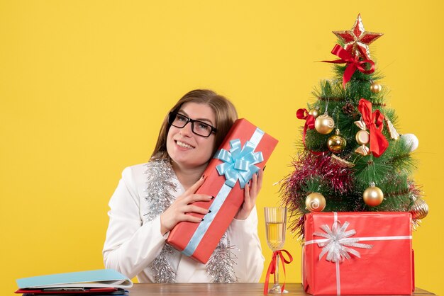 Vooraanzicht vrouwelijke arts zittend met kerstcadeautjes en boom op geel bureau