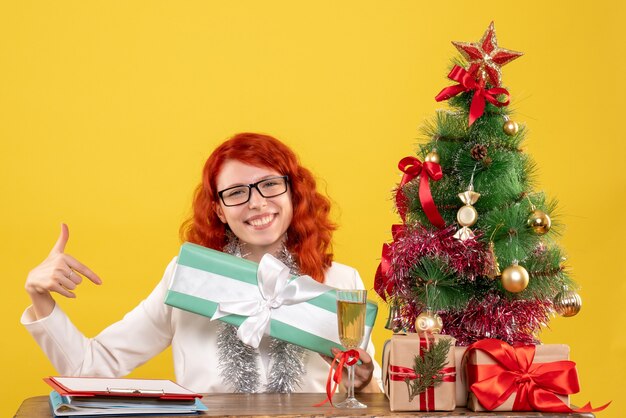 Vooraanzicht vrouwelijke arts zittend met kerstcadeautjes en boom op gele achtergrond