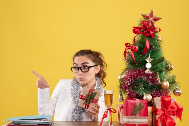 Vooraanzicht vrouwelijke arts zittend met kerstcadeautjes en boom op gele achtergrond