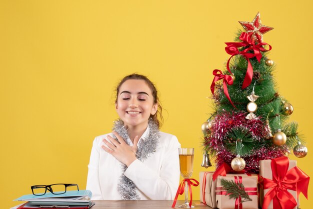 Vooraanzicht vrouwelijke arts zittend rond kerstcadeautjes en boom lachend op gele achtergrond