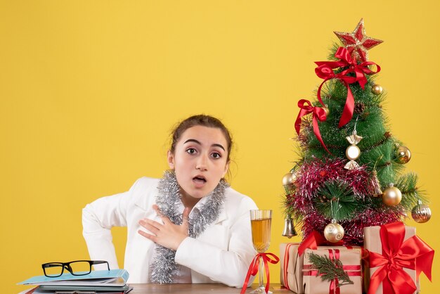 Vooraanzicht vrouwelijke arts zittend rond kerstcadeautjes en boom op gele achtergrond