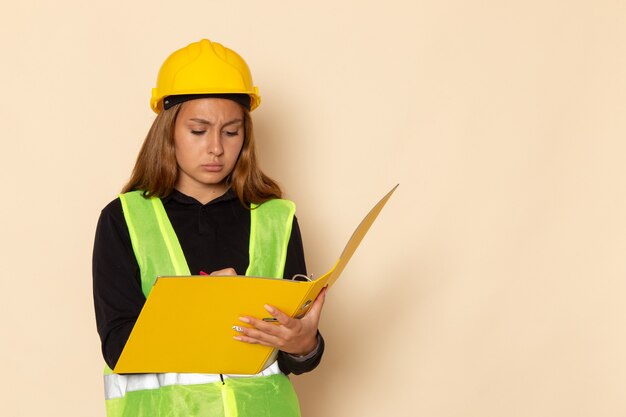 Vooraanzicht vrouwelijke bouwer in gele helm die geel dossier houdt dat nota's op witte muur opschrijft