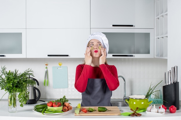 Vooraanzicht vrouwelijke chef-kok in kok hoed die plakjes komkommer op haar gezicht in de keuken zet