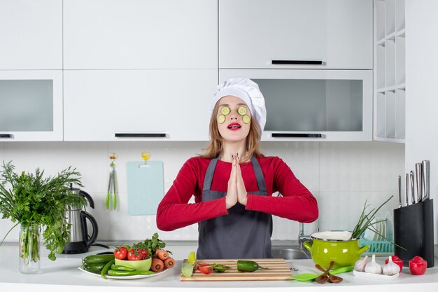 Vooraanzicht vrouwelijke chef-kok in uniform die plakjes komkommer op haar gezicht legt en de handen ineen slaat
