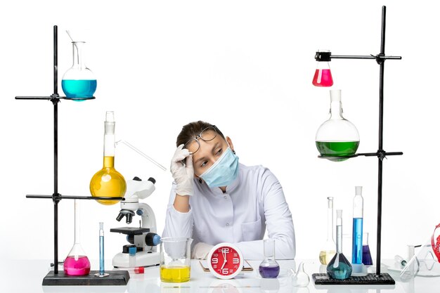 Vooraanzicht vrouwelijke chemicus in medisch pak met masker zittend met oplossingen op witte achtergrond splash virus chemie covid lab