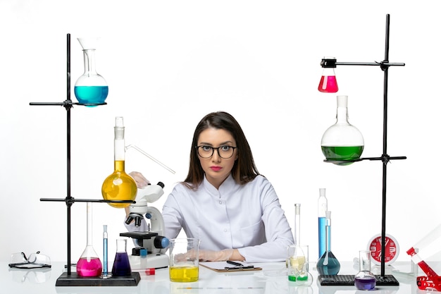 Vooraanzicht vrouwelijke chemicus in wit medisch pak zittend op lichte witte achtergrond lab virus covid pandemie wetenschap