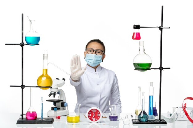 Vooraanzicht vrouwelijke chemicus in witte medische pak met masker zittend met oplossingen op witte achtergrond chemicus lab virus covid-splash