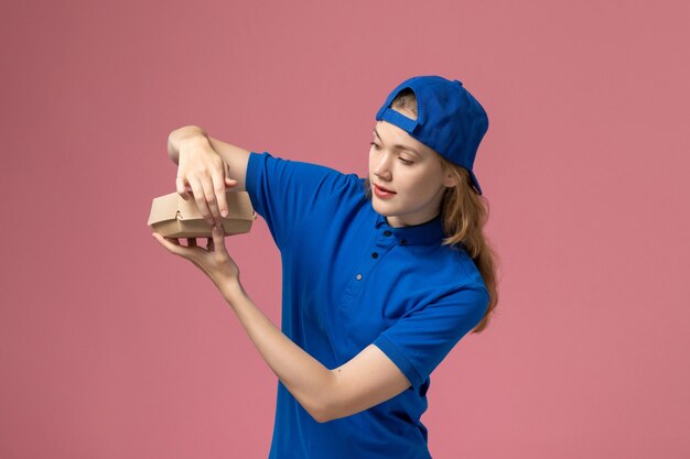 Vooraanzicht vrouwelijke koerier in blauw uniform en cape die een klein voedselpakket voor bezorging vasthoudt en het op de roze muur opent, bezorgdienst uniform