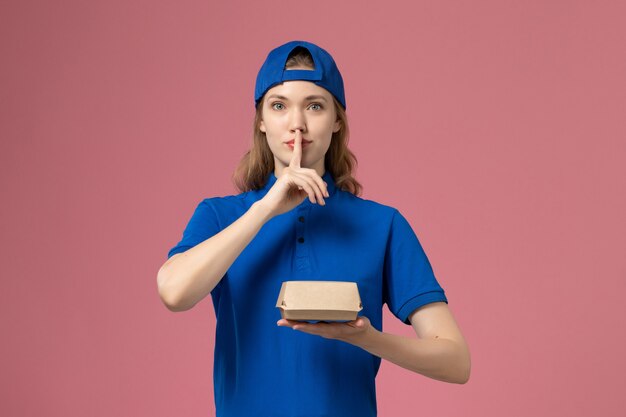 Vooraanzicht vrouwelijke koerier in blauw uniform en cape die weinig bezorgvoedselpakket op de roze muur houdt, bezorgingsuniform dienst werk werknemer meisje baan