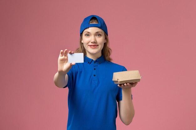 Vooraanzicht vrouwelijke koerier in blauw uniform en cape die weinig voedselpakket met kaart op roze muur, baanbezorgingsmedewerker houdt