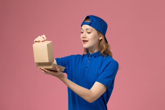 Vooraanzicht vrouwelijke koerier in blauw uniform en cape die weinig voedselpakket voor bezorging op lichtroze muur houdt, baan voor bezorgingsuniform