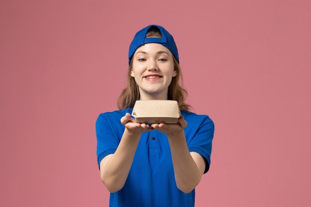 Vooraanzicht vrouwelijke koerier in blauw uniform en cape die weinig voedselpakket voor bezorging op lichtroze muur houdt, uniforme servicemedewerker bezorgwerk