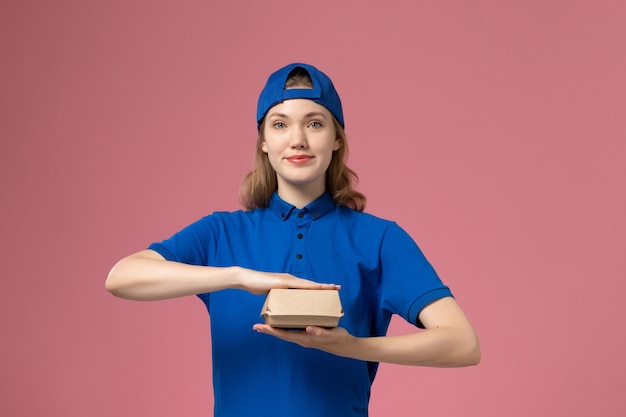 Vooraanzicht vrouwelijke koerier in blauw uniform en cape die weinig voedselpakket voor bezorging op roze muur houdt, werk van een uniform dienstverlenend bedrijf