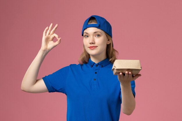 Vooraanzicht vrouwelijke koerier in blauw uniform en cape die weinig voedselpakket voor bezorging op roze muur houdt, werk van een uniform dienstverlenend bedrijf