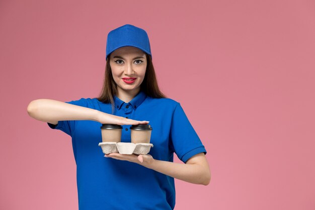 Vooraanzicht vrouwelijke koerier in blauw uniform en cape levering koffiekopjes houden en glimlachend op roze muur