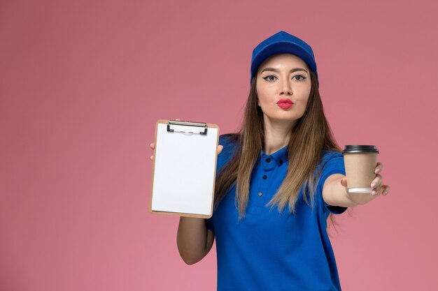 Vooraanzicht vrouwelijke koerier in blauw uniform en cape met bezorging koffiekopje met blocnote op de lichtroze muur