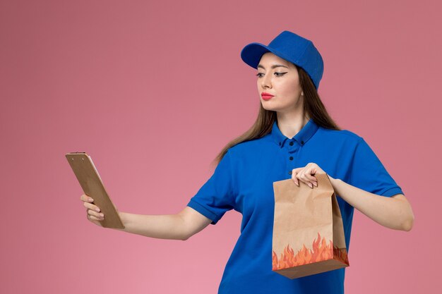 Vooraanzicht vrouwelijke koerier in blauw uniform en cape met blocnote papier voedselpakket op de roze muur