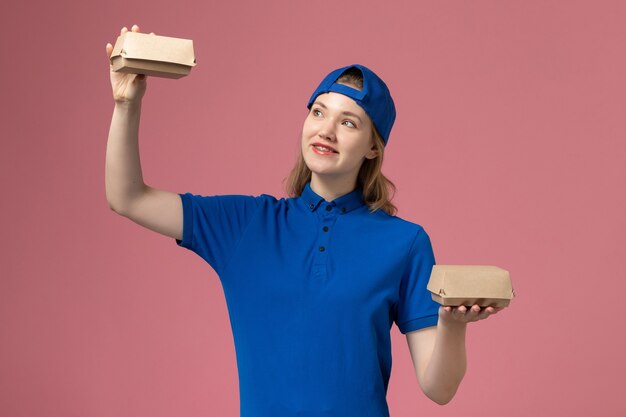 Vooraanzicht vrouwelijke koerier in blauw uniform en cape met kleine bezorgvoedselpakketten op roze muur, bezorgbaan werk uniform servicebedrijf