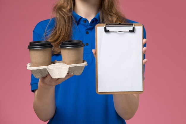 Vooraanzicht vrouwelijke koerier in blauw uniform met bruine bezorgkoffiekopjes en blocnote op de roze bureau-service uniforme bedrijfsbaan