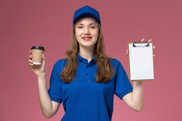 Vooraanzicht vrouwelijke koerier in blauw uniform met bruine koffiekopje met blocnote glimlachend op roze achtergrond service job uniform leverende bedrijf