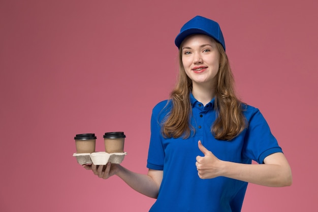 Vooraanzicht vrouwelijke koerier in blauw uniform met bruine koffiekopjes poseren en glimlachend op de roze achtergrond service uniform bedrijf baan leveren