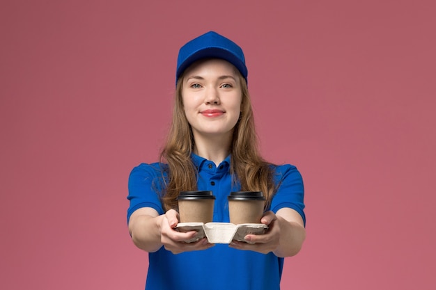 Vooraanzicht vrouwelijke koerier in blauw uniform met bruine levering koffiekopjes glimlachend op roze achtergrond service uniforme bedrijfsmedewerker