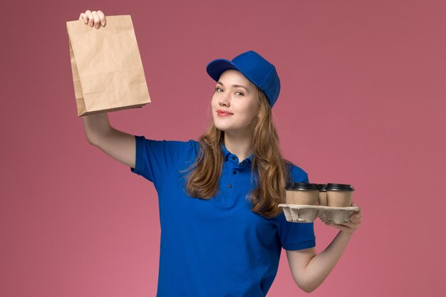 Vooraanzicht vrouwelijke koerier in blauw uniform met bruine levering koffiekopjes met voedselpakket glimlachend op roze bureau service uniforme bedrijfsmedewerker
