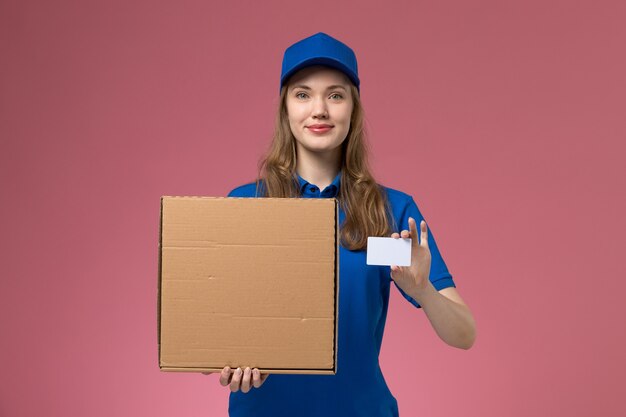 Vooraanzicht vrouwelijke koerier in blauw uniform met voedselleveringsdoos en witte kaart op de roze werknemer van het bureau dienstuniform