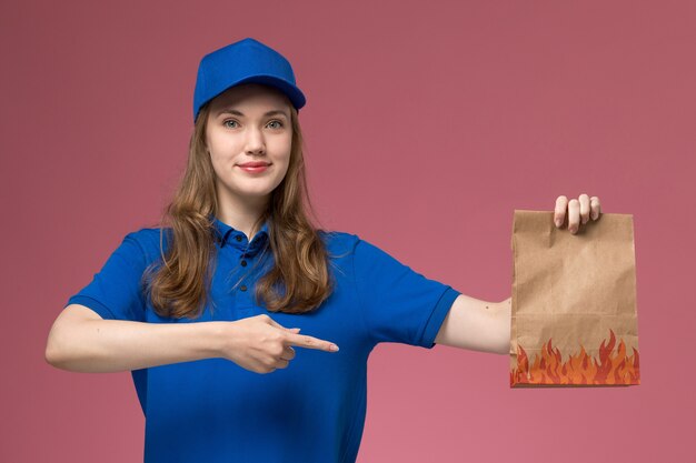 Vooraanzicht vrouwelijke koerier in blauw uniform voedselpakket met lichte glimlach op lichtroze bureau baan werknemer dienst uniform bedrijf