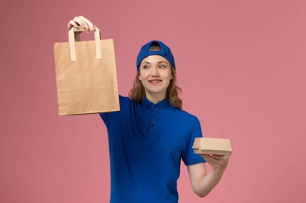 Vooraanzicht vrouwelijke koerier in blauwe uniforme cape met bezorgpakketten op de roze muur, werk van de dienstverlening van de werknemer