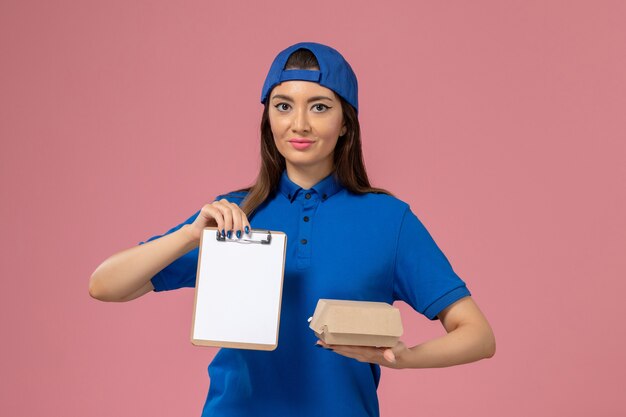 Vooraanzicht vrouwelijke koerier in blauwe uniforme cape met klein afleverpakket met blocnote op lichtroze muur, levering van de baan van het dienstmeisje