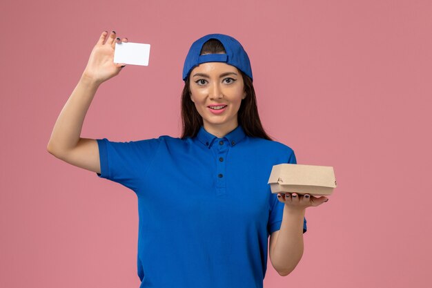 Vooraanzicht vrouwelijke koerier in blauwe uniforme cape met klein afleverpakket met plastic kaart op lichtroze muur, levering van werknemersdienst