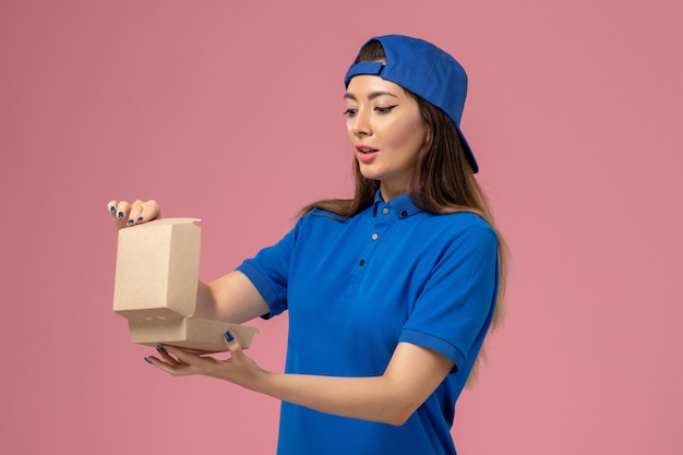 Vooraanzicht vrouwelijke koerier in blauwe uniforme cape met klein afleverpakket openen op roze muur, levering door werknemer