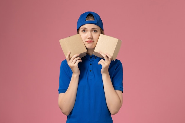 Vooraanzicht vrouwelijke koerier in blauwe uniforme cape met kleine pakketjes van het bezorgvoedsel op roze muur, medewerker van de bezorgdienst