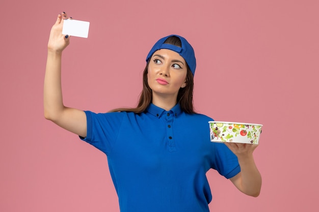 Vooraanzicht vrouwelijke koerier in blauwe uniforme cape met leveringskom met kaart op lichtroze muur, servicemedewerker bezorger