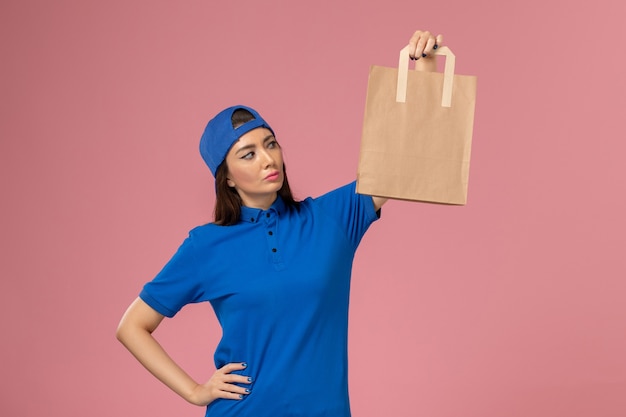 Vooraanzicht vrouwelijke koerier in blauwe uniforme cape met papieren bezorgpakket op lichtroze muur, servicemedewerker werk leveren